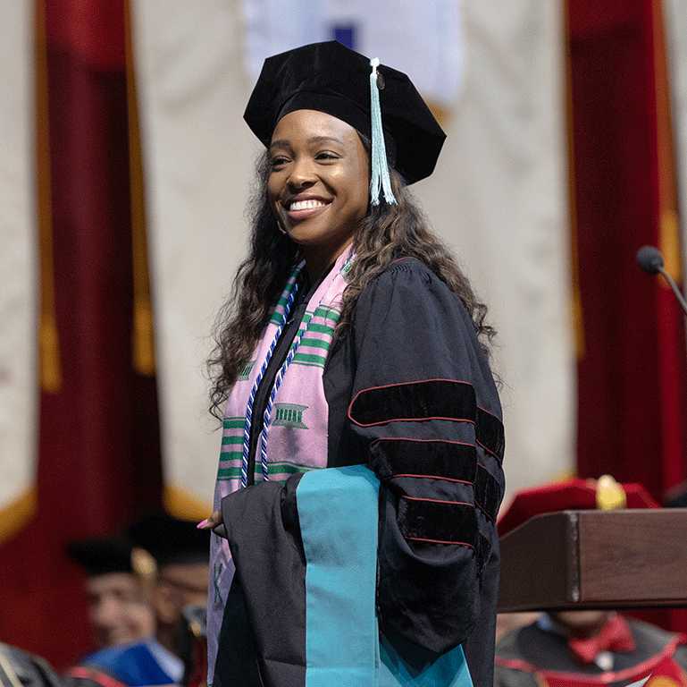 A female doctoral graduate smiles as she holds her doctoral hood over her arm at commencement.