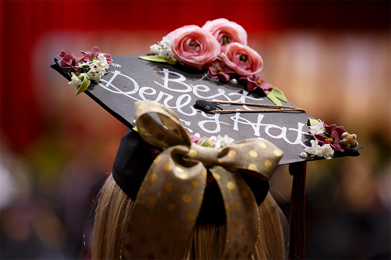 A young woman sports a lavishly decorated mortarboard proclaiming in cursive type "best derby hat," a reference to the ostentatious millinery typically worn by women attending the United State's annual Kentucky Derby horse race. 