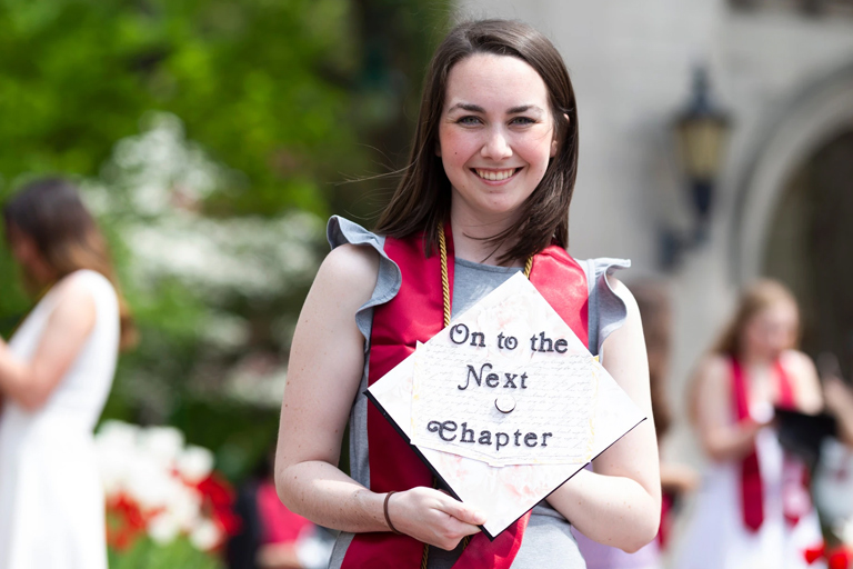 A young woman in a summer dress stands at the Sample Gates with her decorated graduation cap.