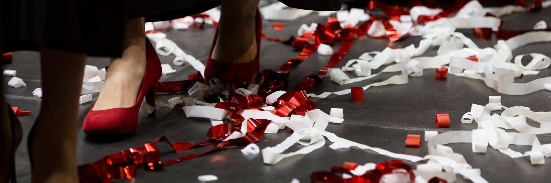 An ankle view of a young woman wearing red heels picking her way through a heap of red and white confetti.