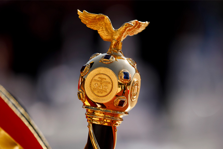 A shallow-focus image of the official mace of Indiana University, a jewel-encrusted golden orb stamped with greek lettering on all four sides and capped with a sculpture of an eagle taking flight. 