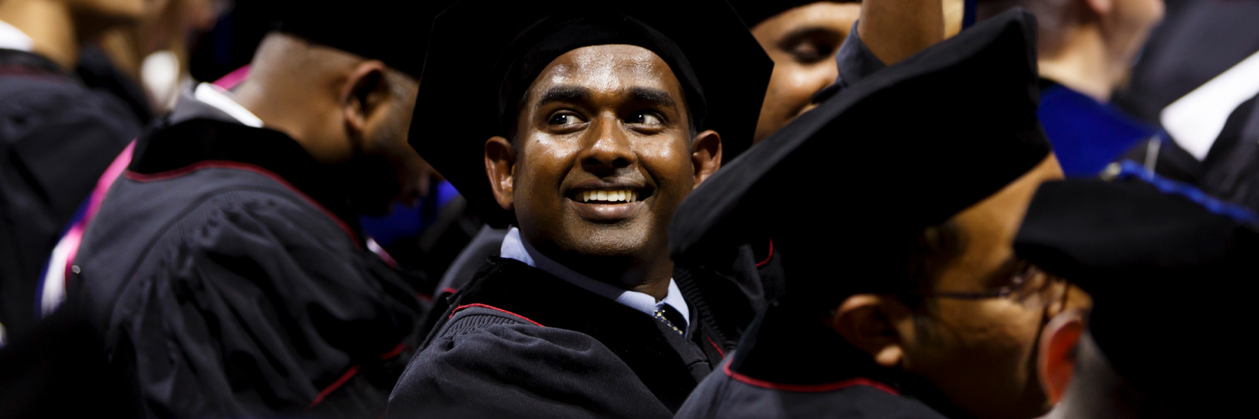 A male masters' or doctoral student gazes upward in awe, his face the only identifiable one in a sea of dark robes and caps. 