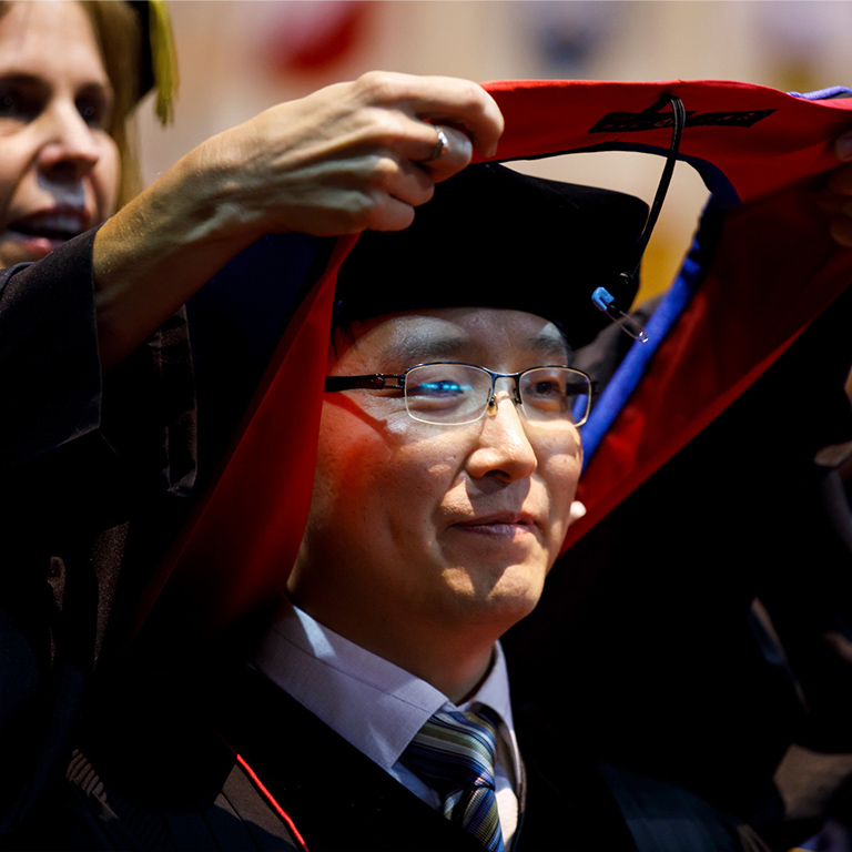 A male doctoral student in glasses tears up as he is hooded during the graduate commencement ceremony. 