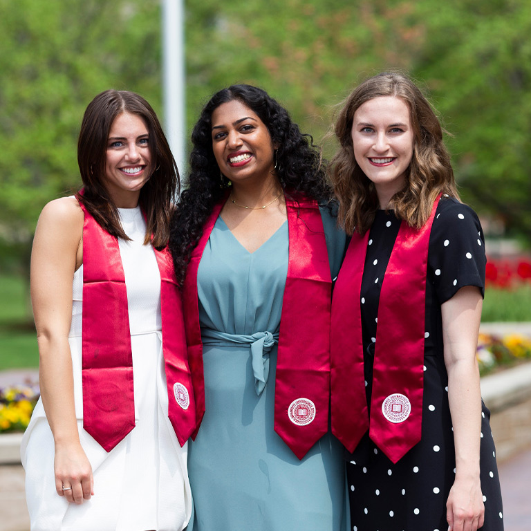Three young women in spring dresses and crimson Stoles of Gratitude gather together for a photo by the Sample Gates. 