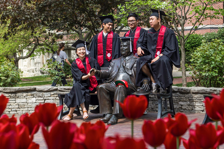 A group of male and female students in graduation attire crowd around the bronze statue of former university president Herman B Wells for a commemorative photo.