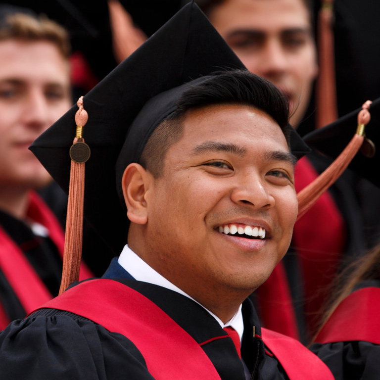 A young man in a crisp white shirt, red tie, and graduation attire smiles towards the camera at Commencement. 