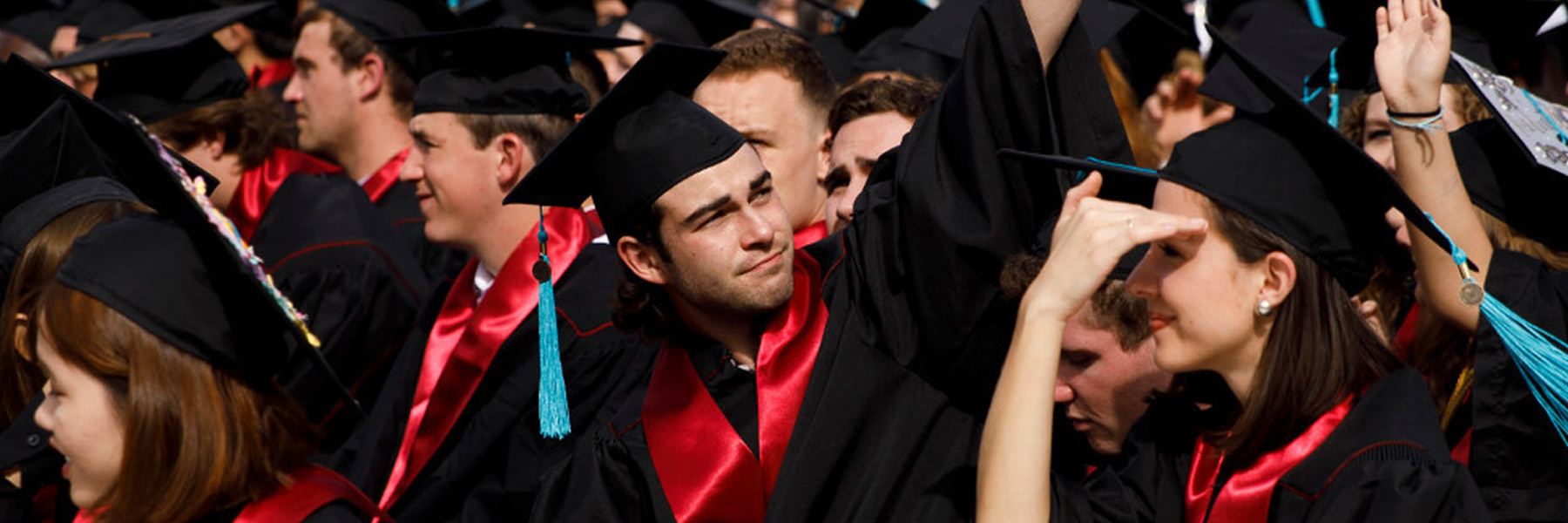 A young man in a crowd of graduates raises his hand triumphantly and looks upward. 