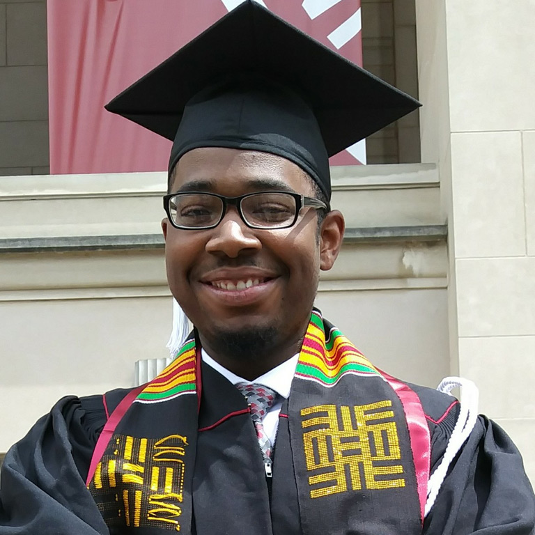 A young man proudly wearing his graduation attire and Neal Marshall Black Culture Center kente stole poses in front of IU Auditorium on IU Bloomington's campus. 