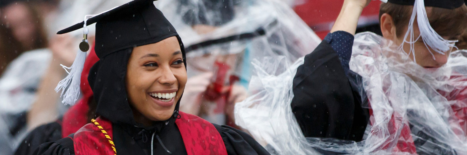 A young woman in graduation attire and modestly covered hair smiles in the middle of a light rain. In the background, other soon to be graduates struggle to put on rain ponchos.