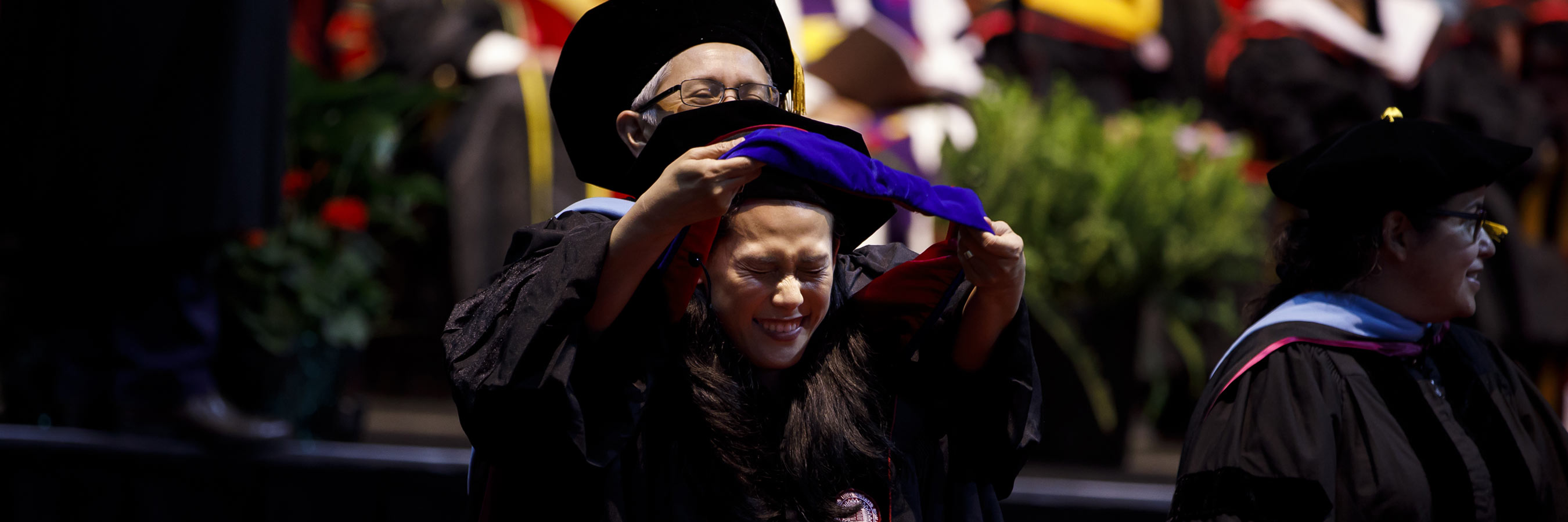 A female graduate student scrunches up her face in excitement as she is hooded by a smiling faculty member. 