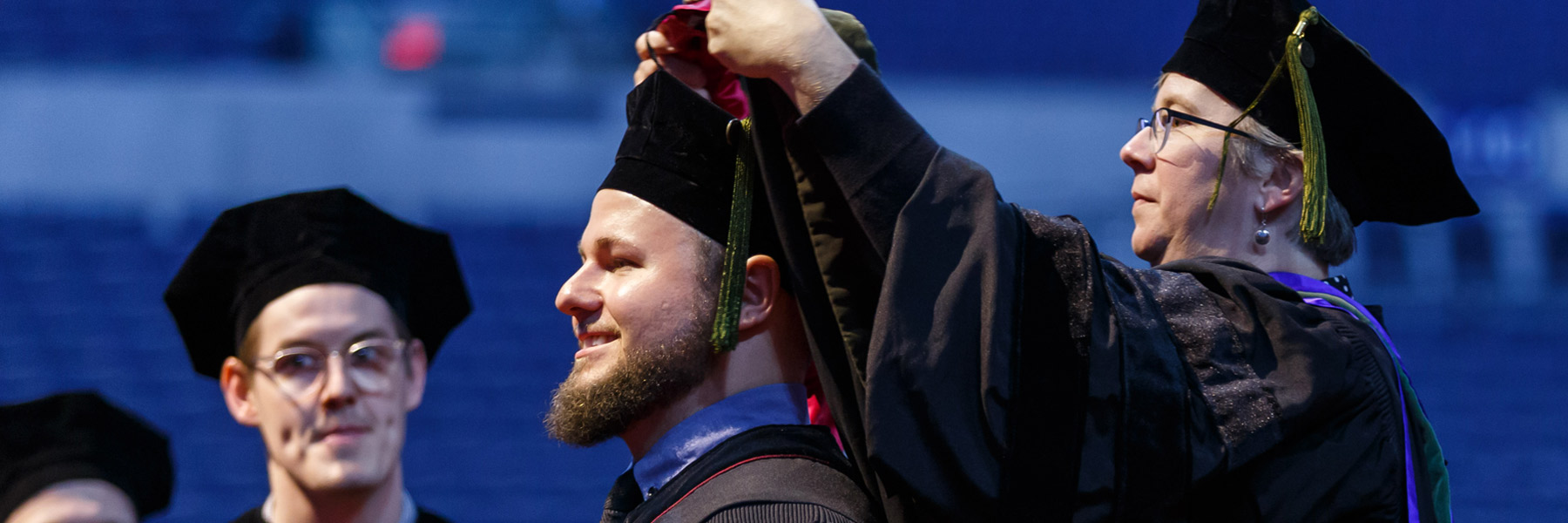 A dramatic blue-toned image of a middle-aged female professor hooding a smiling, bearded young male student at commencement.