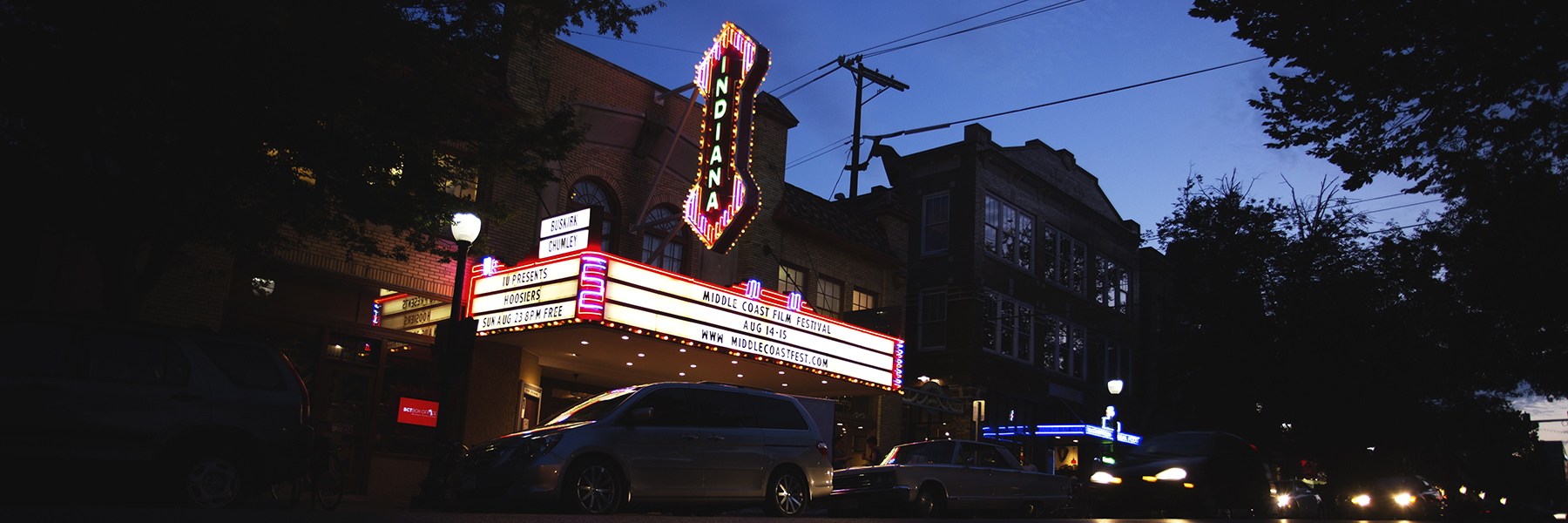 A glorious photo of the Buskirk-Chumley's neon marquee after dark, silhouetted by near-black trees. In front of the theatre, cars speed by against the night sky.