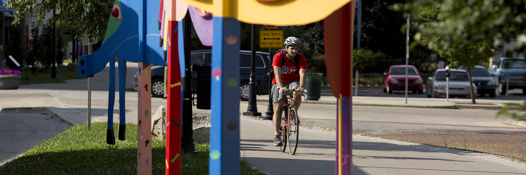 A male cyclist travels by brightly colored sculptural art on Bloomington's B-line trail in summer. 
