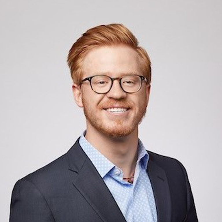 A professional headshot of a bearded young man with glasses and light red hair, wearing business casual dress.