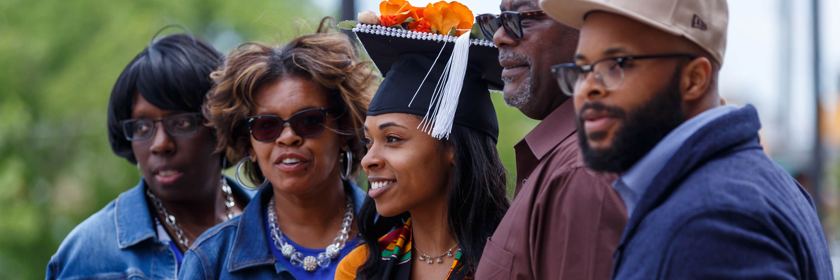 A graduate wearing a mortarboard decorated with bright orange flowers smiles for the camera. She is flanked by a group of relatives in casual dress; two older women on one side, two younger men on the other. 