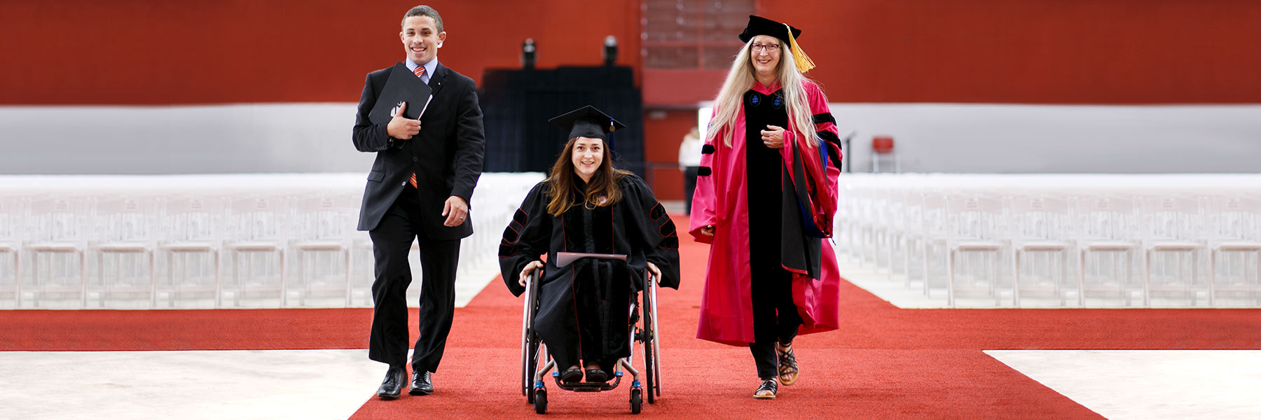 A young graduate in a wheelchair and her graduation attire moves down the red carpet. She is flanked by a smiling man and a female faculty member in full academic regalia.