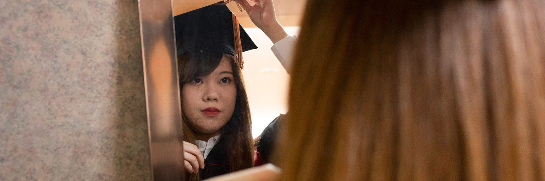 A young woman adjusts her mortarboard cap in a mirror at GradFair